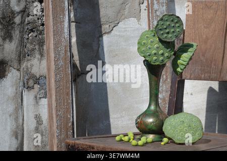 A slender, antique bronze vase rests on a weathered wooden surface, displaying several vibrant green lotus seed pods. A handful of small, round seeds Stock Photo