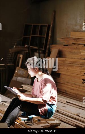 Asian female carpenter writing on clipboard while sitting sideways on stack of wooden planks in workshop on sunny day Stock Photo