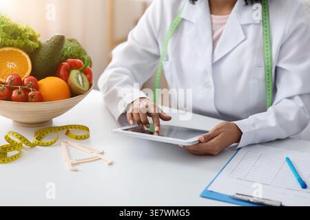 Cropped image of Black female dietitian with tape measure around her neck using tablet while sitting at table with bowl of fruits and vegetables in... Stock Photo