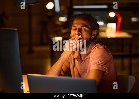 Indian male programmer yawning while sitting at table with laptop working late at night in office Stock Photo
