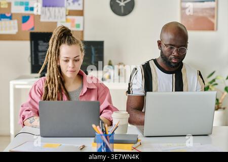 Black man and caucasian woman working on laptops while sitting together at table in office Stock Photo