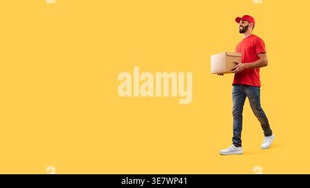 Arab delivery man wearing cap holding cardboard box walking against yellow background in studio Stock Photo