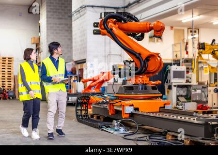 Asian man and caucasian woman in green safety vests checking robotic arms in factory Stock Photo