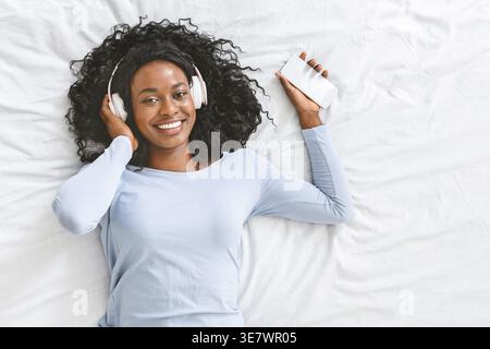 Smiling Black woman with curly hair listening to music with headphones holding smartphone lying in bed at home Stock Photo