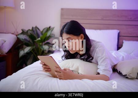Serious Asian woman using tablet lying on bed at home Stock Photo