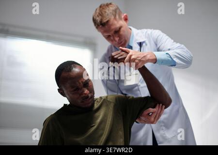 Black military man sitting with white male doctor examining his arm standing behind him in hospital Stock Photo