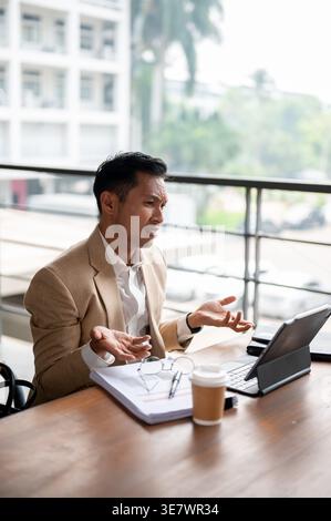 Disappointed Asian businesman shrugging shoulders having online meeting on tablet sitting at wooden table with documents at coffee on balcony in hi... Stock Photo