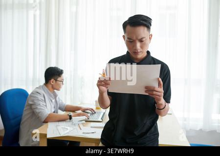 Disappointed Asian engineer checking project plan standing near male colleague working on laptop in office Stock Photo