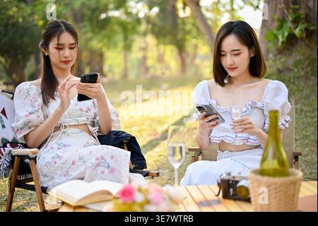 Asian female friends using smartphones while sitting together at table with books and camera having picnic in park during daytime Stock Photo