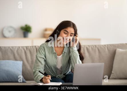 Indian female in headset writing down notes having online class on laptop sitting on sofa at home Stock Photo