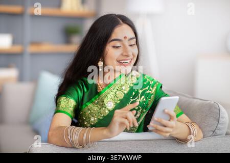 Smiling Indian female in traditional sari dress using smartphone sitting on sofa at home Stock Photo