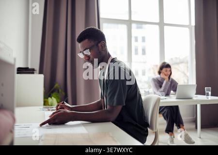 Serious Black male tailor wearing eyeglasses reviewing new fashion designs while sitting at table near caucasian tailor in studio Stock Photo