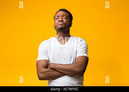 Black man in white shirt pouting lips standing with arms crossed against yellow background in studio Stock Photo