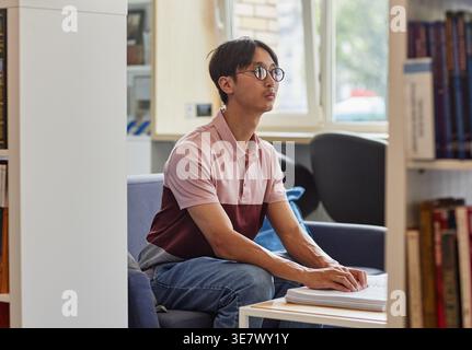 Blind Asian male student wearing eyeglasses reading book in tactile braille sitting on couch in college library during daytime Stock Photo