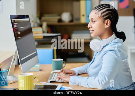 Mixed race female programmer with headphones around her neck writing codes for program on computer while sitting at desk in office Stock Photo