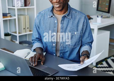 Smiling Black man wearing denim long sleeves analyzing documents and working on laptop while sitting at desk in office Stock Photo
