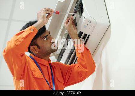 Indian male technician in orange bodysuit using screwdriver while setting up air conditioner in costumer's home Stock Photo