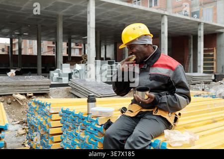 Black man worker eating sandwich holding cup while sitting on stack of building materials in construction site during daytime Stock Photo