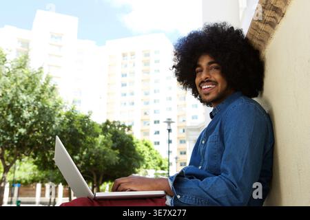 Smiling Black man with afro hairstyle working on laptop sitting leaning on wall outdoors on sunny day Stock Photo