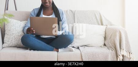 Cropped image of Black woman with twists working on laptop sitting cross-legged on couch in the living room at home Stock Photo