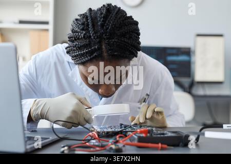 Focused Black female computer engineer with braids using magnifying glass examining electronic parts sitting at table in quality control lab Stock Photo