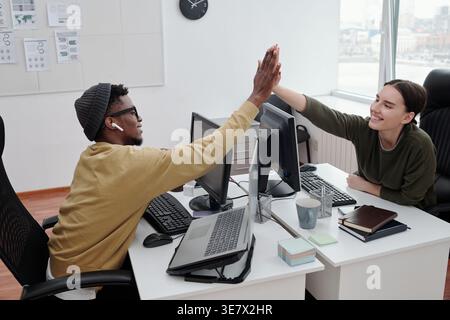 Smiling Black man and Mixed race woman doing high five working together sitting at table in office Stock Photo