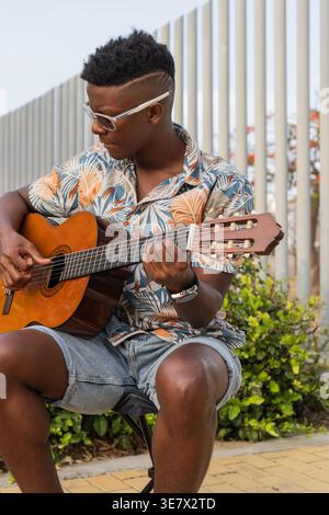 Hispanic man wearing glasses playing guitar sitting on stool against lush foliage garden during sunset Stock Photo