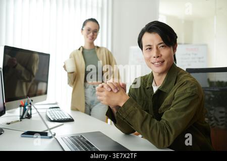 Asian male programmer sitting with hands together at table with laptop near female colleague in office Stock Photo