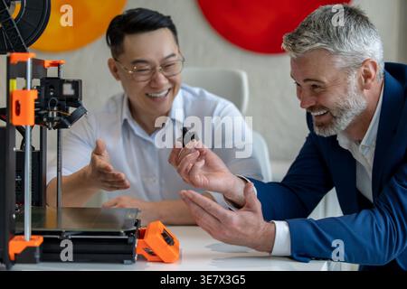 Asian and caucasian male industrial engineers examining product created by 3D printer while sitting at table working together in office Stock Photo