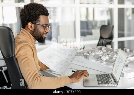 African American male architect wearing eyeglasses reviewing blueprints while using laptop in office Stock Photo