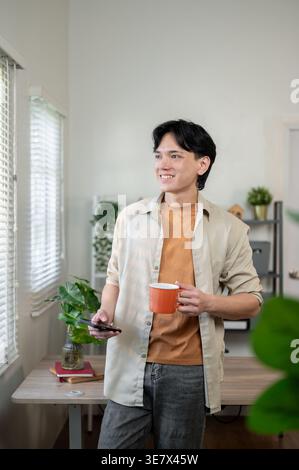 Thai businessman holding smartphone and cup of coffee looking away while standing and leaning against table in office Stock Photo