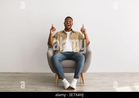 Emotional Black Guy Pointing Fingers Upward Looking Up, Advertising Great Idea Sitting In Comfortable Chair Over Gray Wall At Home. Smiling To Camera. Stock Photo