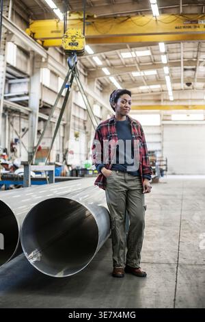 Black woman standing with hand on pocket near aluminum pipes in factory Stock Photo