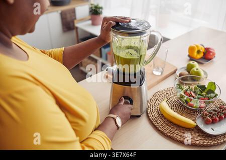Plus sized black woman using blender making green smoothie with fruits standing at table in kitchen at home Stock Photo
