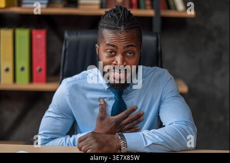 Black businessman with hand on chest sitting at table in office Stock Photo
