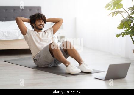 Asian man with curly hair working on abs watching fitness video on laptop while sitting on yoga mat in bedroom at home Stock Photo