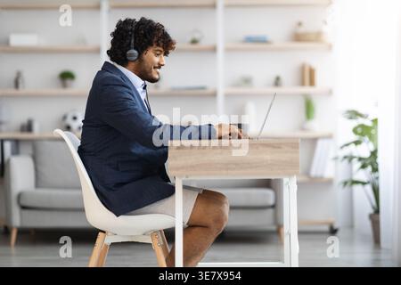 Asian man in headset wearing suit shorts having video call using laptop while sitting at table in living room at home during work from home Stock Photo