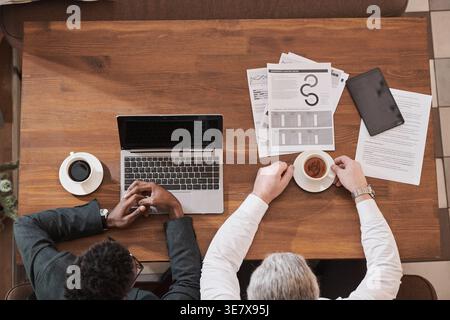 Black and caucasian businessmen sitting at table with coffee mugs, laptop, and tablet during lunch meeting in restaurant Stock Photo