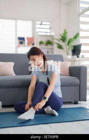 Adult Asian woman happily prepares her indoor exercise session. She adjusts socks on a blue yoga mat in a bright spacious home. Focus on personal heal Stock Photo