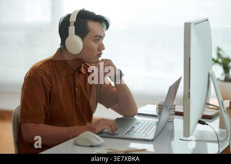 Asian male software developer wearing headphones testing new software on laptop sitting at desk in office Stock Photo