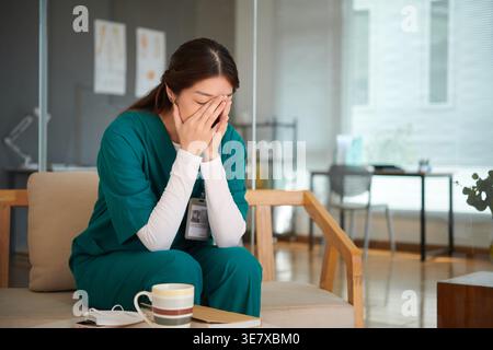 Asian female nurse with hands covering face wearing scrub suit sitting on sofa near table with book and coffee cup in hospital lounge Stock Photo