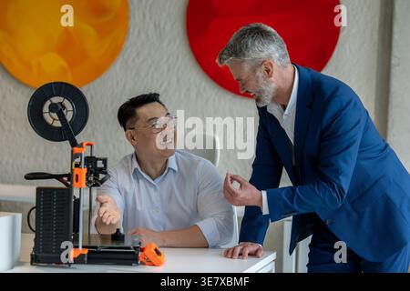 Asian male industrial engineer wearing eyeglasses sitting at table and presenting 3D printer to caucasian male colleague standing beside him in office Stock Photo