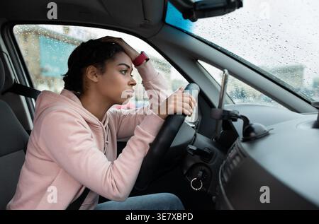 Black female driver looking away touching head leaning on steering wheel while sitting inside car during rainy day Stock Photo