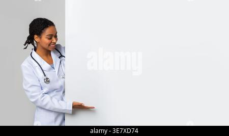 Free place for your ad. black female doctor looking and pointing at big blank advertising board with open palm, empty space for text, panorama Stock Photo