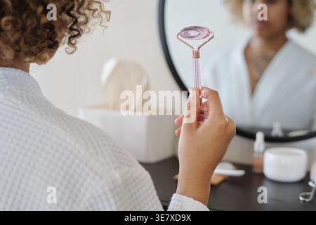 Black woman in bathrobe holding facial roller standing in front of mirror in bathroom Stock Photo