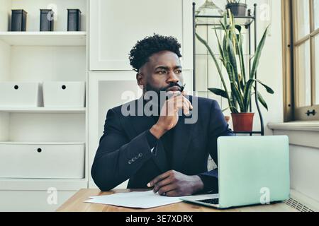 Black man with beard holding pen looking away thinking while sitting at table with laptop and papers near window in office Stock Photo