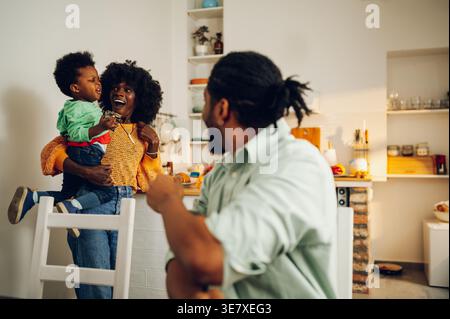 Cheerful Black mother with afro holding her son standing in kitchen near husband sitting at table and looking at them at home Stock Photo