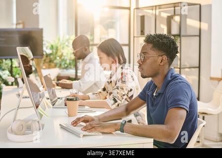 Black male programmer with afro wearing eyeglasses working on computer while sitting at desk beside colleagues in office Stock Photo