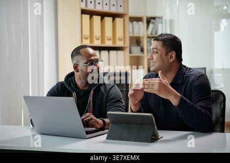 Asian male software developer and project manager discussing new product while sitting at table with tablet and laptop in office Stock Photo