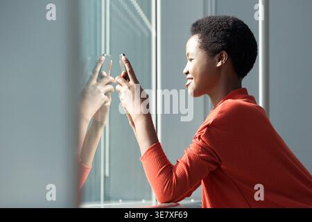 Smiling Black woman reading phone messages while leaning on window pane in airport terminal Stock Photo
